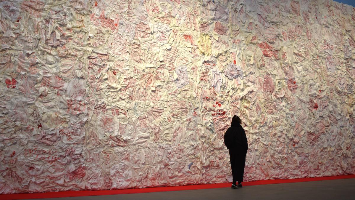 Eine Person steht vor einer riesigen Wand aus 690 weißen Blusen, auf die Frauen mit rotem Stift ihre Geschichte geschrieben haben. Die Wand ist mindestens dreimal so hoch wie die Person.