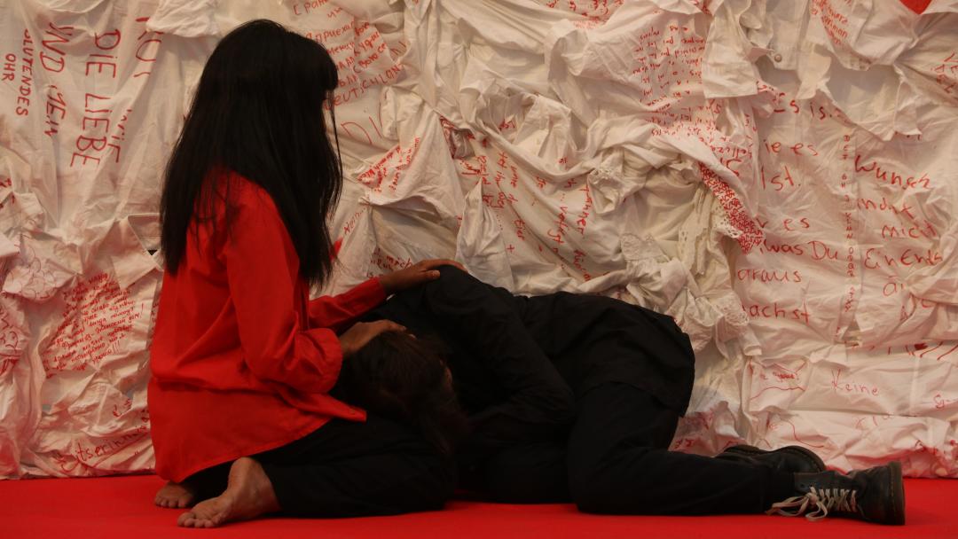 Die Tänzerin Maira Aldana Baeza in roter Bluse kniet vor der Kunstinstallation, die Sängerin liegt in ihrem Schoß.